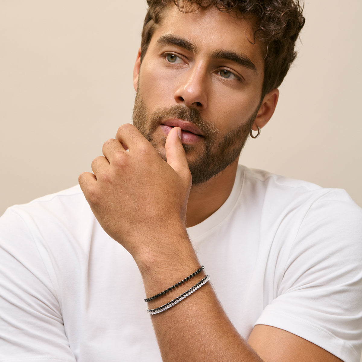 Man wearing white t-shirt with stylish bracelets, model posing with thoughtful expression, showcasing accessories from fashion collection.