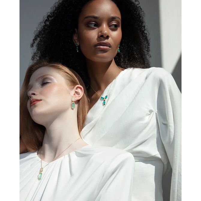 Two women wearing elegant white dresses and emerald jewelry under soft lighting.