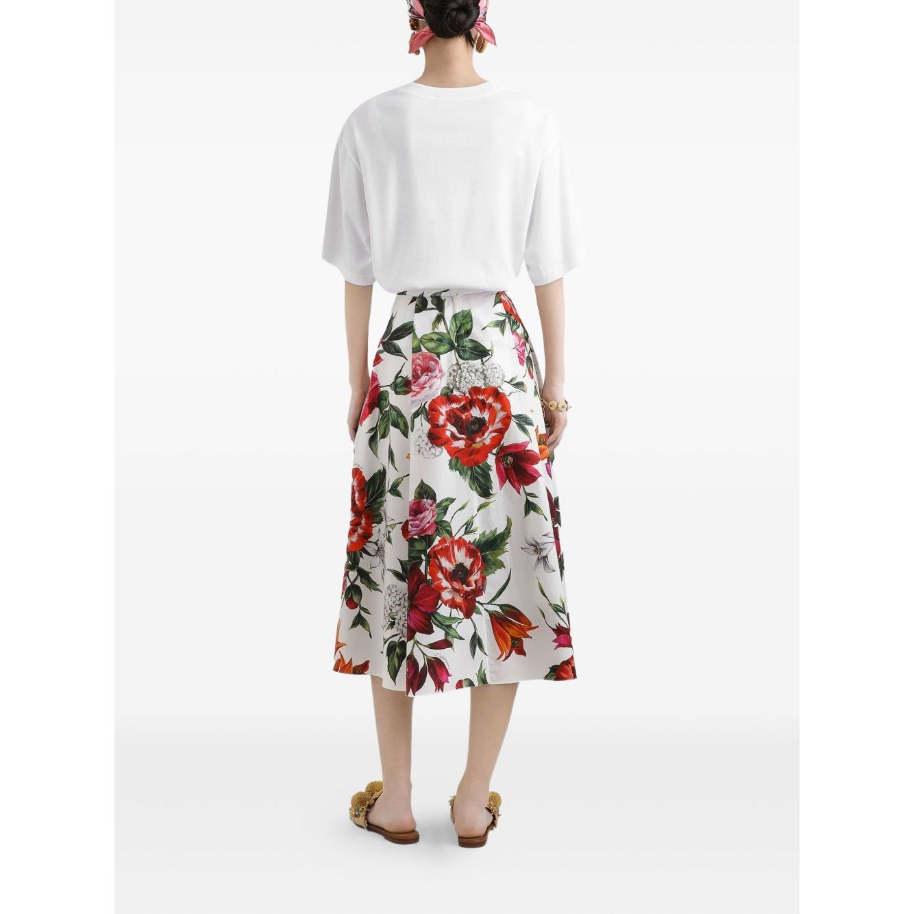 Back view of woman wearing white shirt and floral midi skirt with red and green pattern, holding hands by side, standing against white background.