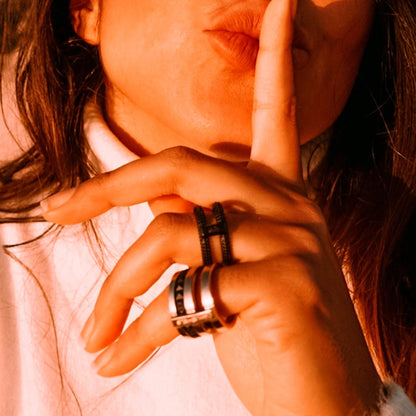 Woman wearing AN JEWELS JEWELRY Mod. AR.R2NS02BK-7 rings, holding finger to lips, close-up on hand in warm lighting.