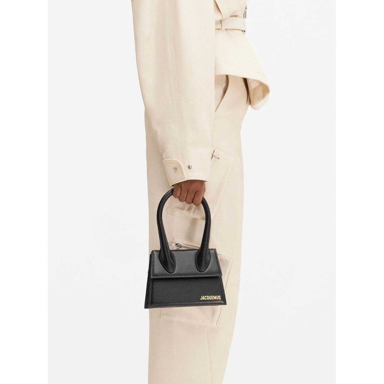 Side view of Jacquemus Le Chiquito Moyen Black Bag with gold logo, held by model in beige outfit, featuring handle and magnetic flap closure.